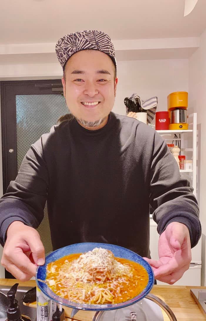 料理を盛った青い皿を持ち、笑顔を見せる鈴木ケンジ